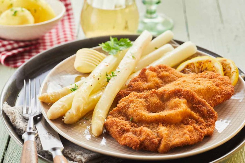Teller mit in zerlassener Butter servierten weißen Spargelstangen, daneben ein knusprig paniertes Schnitzel, Butterflocke und Zitronenscheibe.